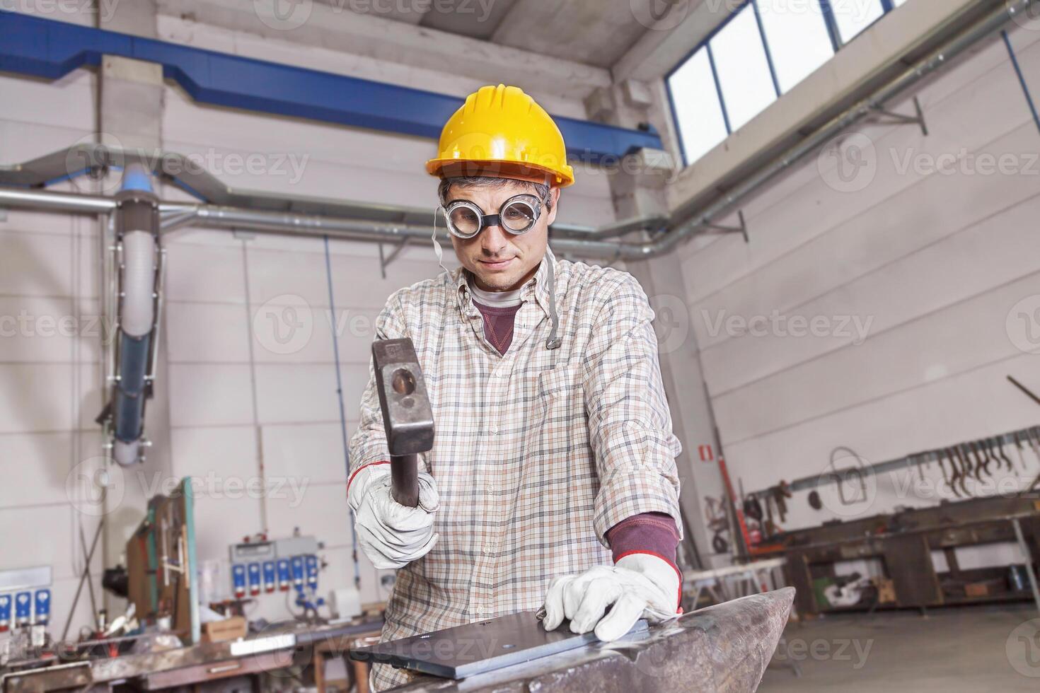 a blacksmith in work clothes with helmet and protective glasses working with hammer and anvil photo