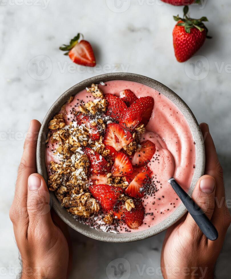 A delicious thick strawberry smoothie bowl topped with fresh sliced strawberries, crunchy granola, and black chia seeds. The bowl is held in two hands photo