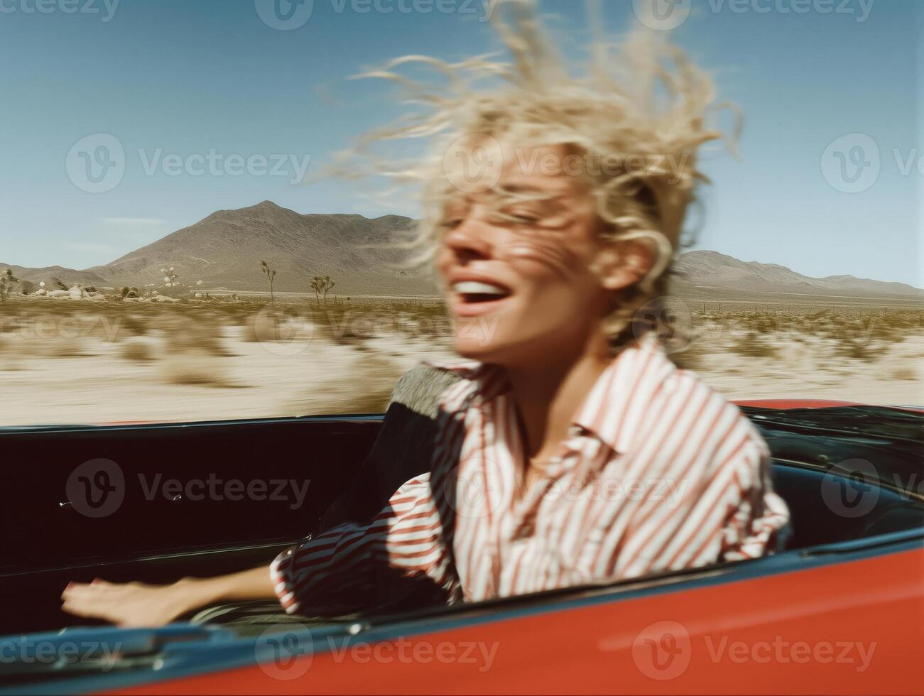 A joyful young blonde woman with wind-blown hair, laughing while riding in a red convertible through a desert landscape. She wears a striped button-up shirt, and her hair is messy from the speed. photo