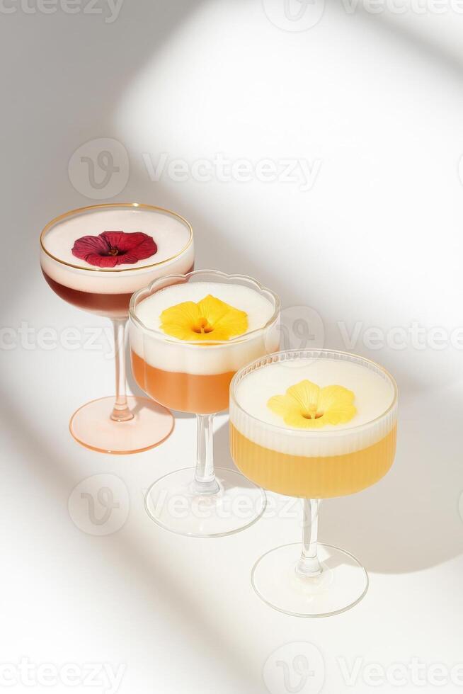 Three cocktails with foam, each topped with an edible flower, served in elegant pastel glasses with varied shapes one with a gold rim, one with scalloped edges, and one ribbed. photo