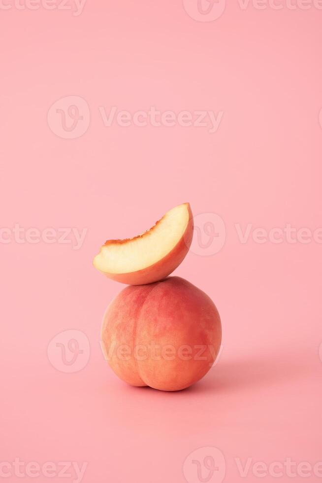 Minimalist food composition showing a whole ripe peach with a fresh slice balanced on top, placed against a soft matte pink background photo