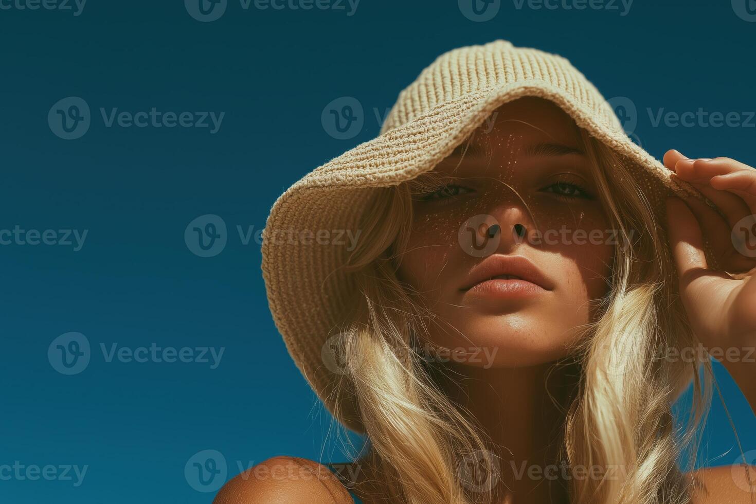 Portrait of a young blonde woman wearing a light woven sun hat, set against a vivid deep blue sky. photo