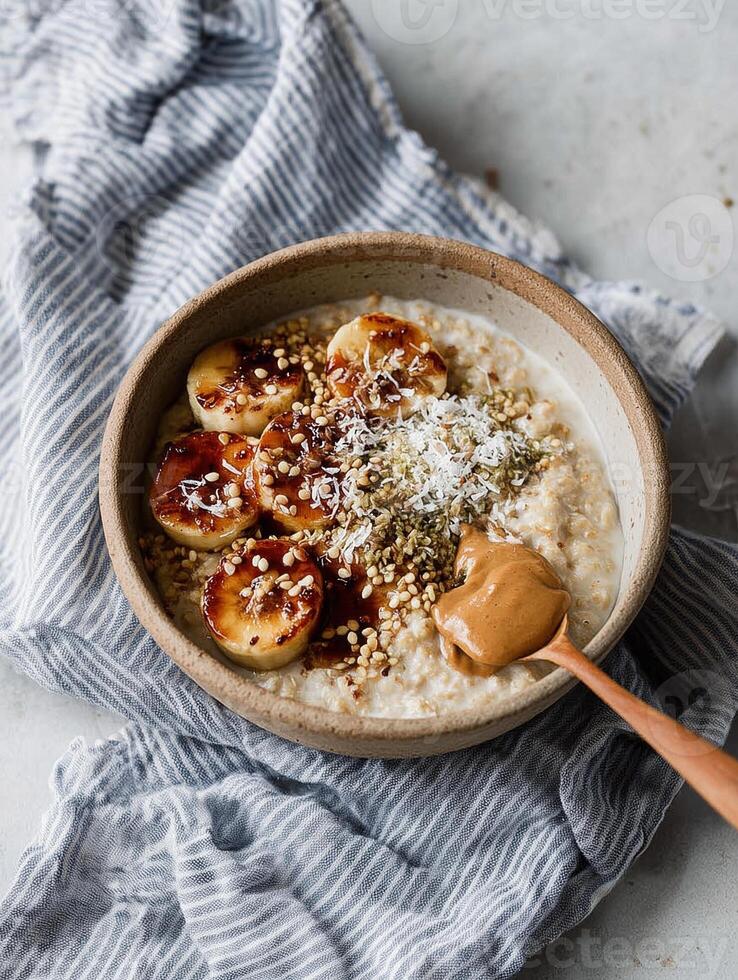 A ceramic bowl filled with creamy oatmeal, topped with caramelized banana slices, hazelnuts, hemp seeds, and a generous spoonful of almond or peanut butter. photo
