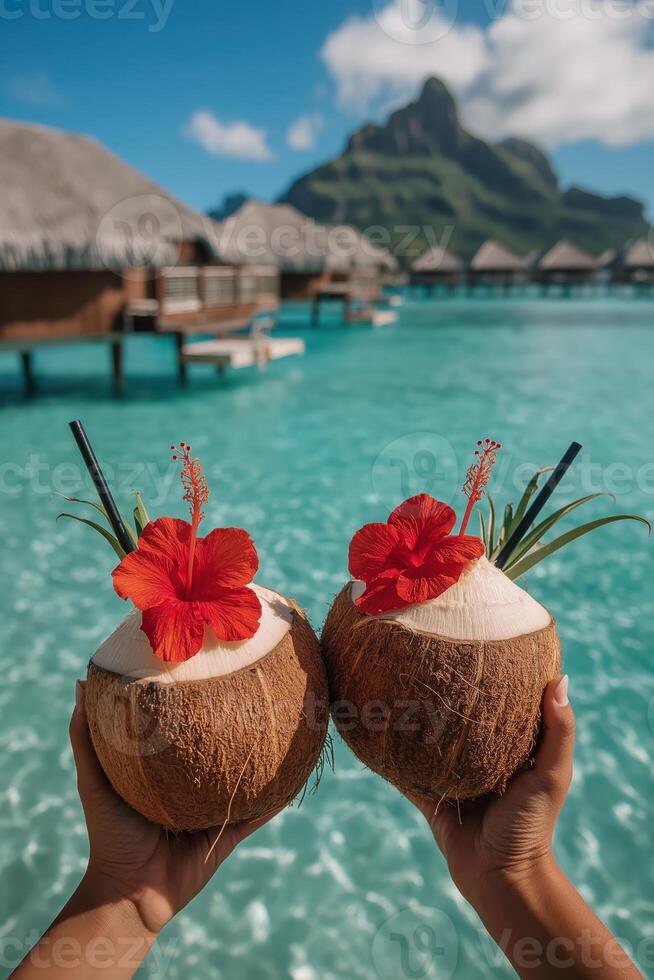 A pair of hands holding two fresh coconuts, each topped with a vivid red hibiscus flower, black straw, and a tropical plant leaf garnish photo