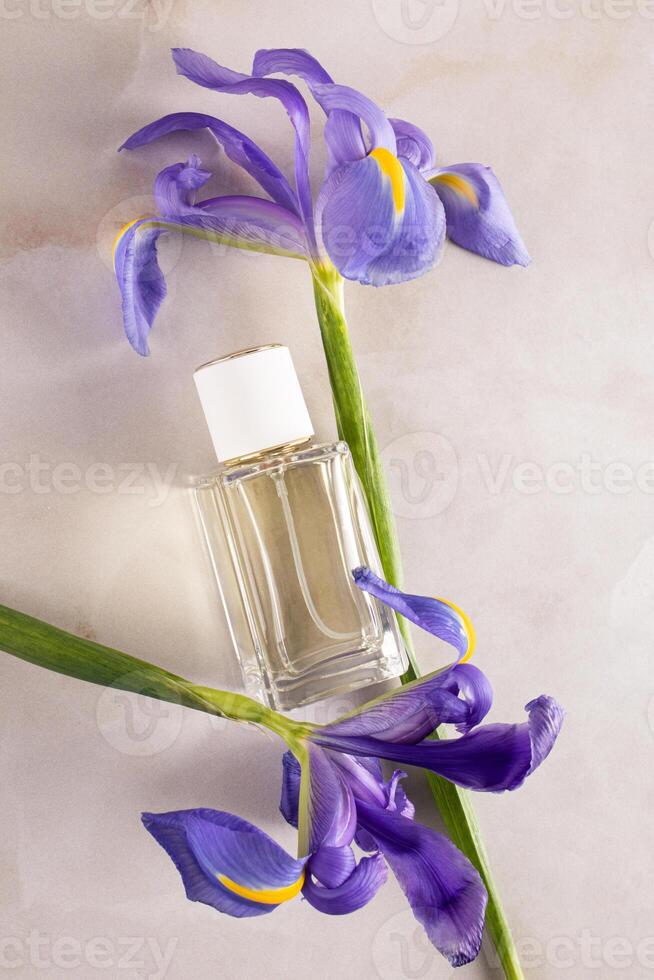 A transparent bottle of perfume on beige marble background among delicate blue irises. The top vertical view. A blank mockup of a non-branded bottle. photo