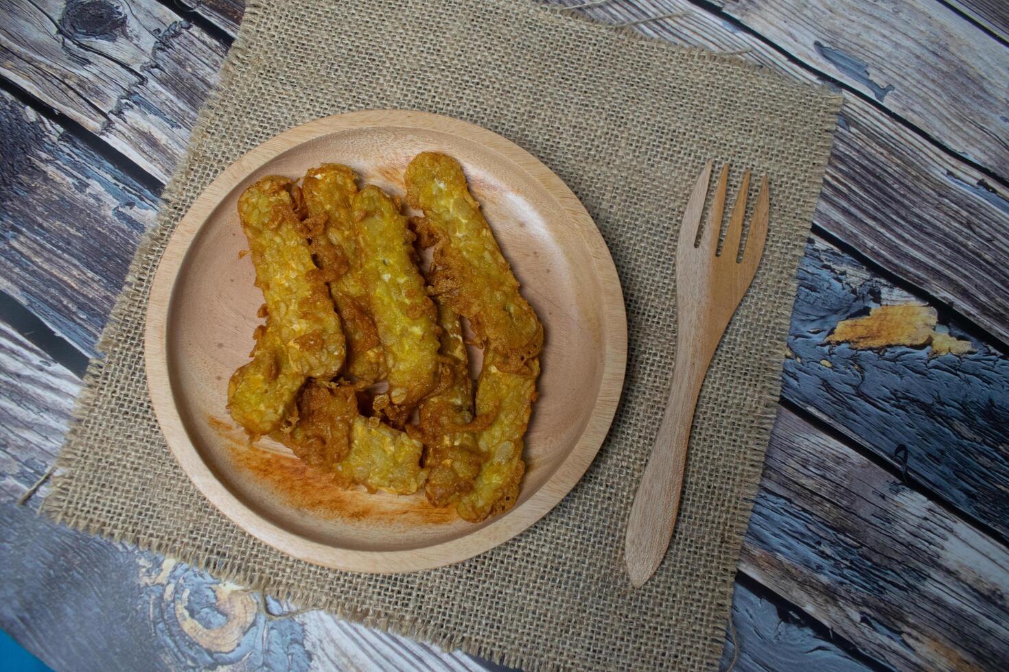 A wooden plate containing flour fried tempeh is served on a burlap cloth on a wooden background photo