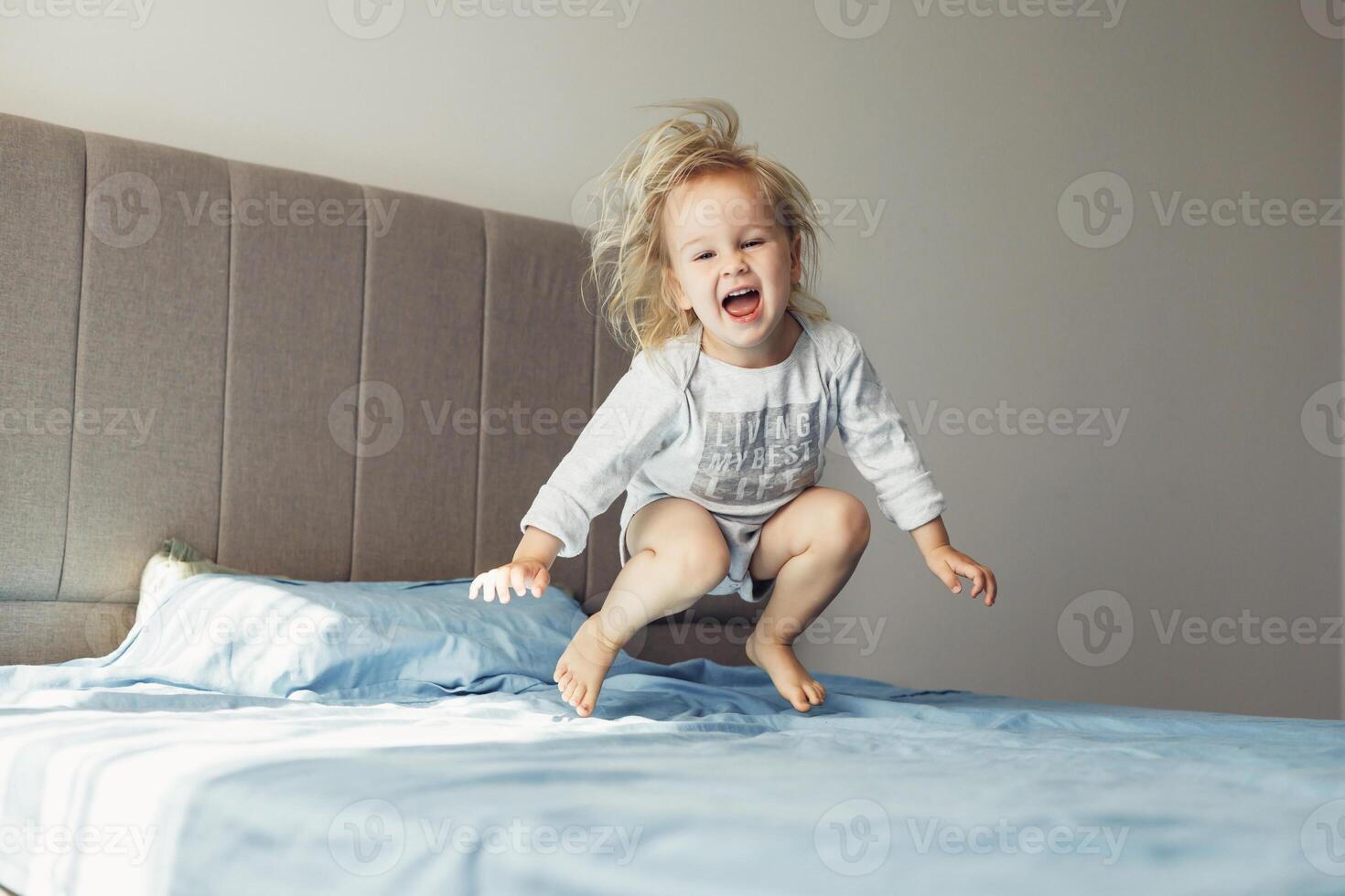 Blonde toddler joyfully jumping on bed at home during sunny playtime photo