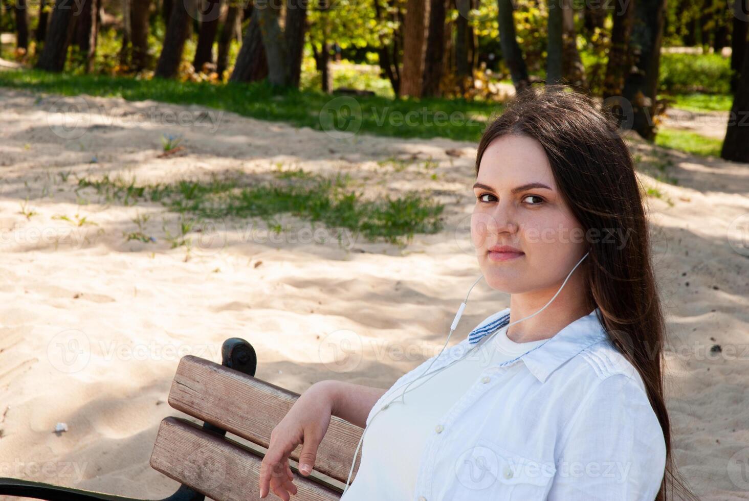 Young caucasian female relaxing on beach bench with headphones, enjoying sunny day in nature, surrounded by sand and trees. Digital nomad lifestyle. Remote work photo