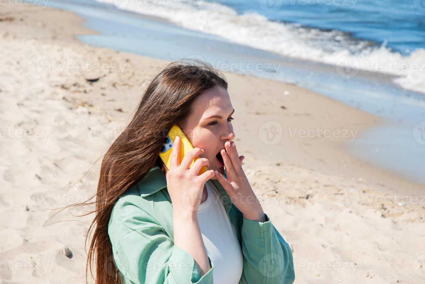 Young caucasian woman on sunny beach making a surprised phone call with a bright yellow smartphone as ocean waves gently lap the shore. Digital nomad lifestyle. Remote work photo