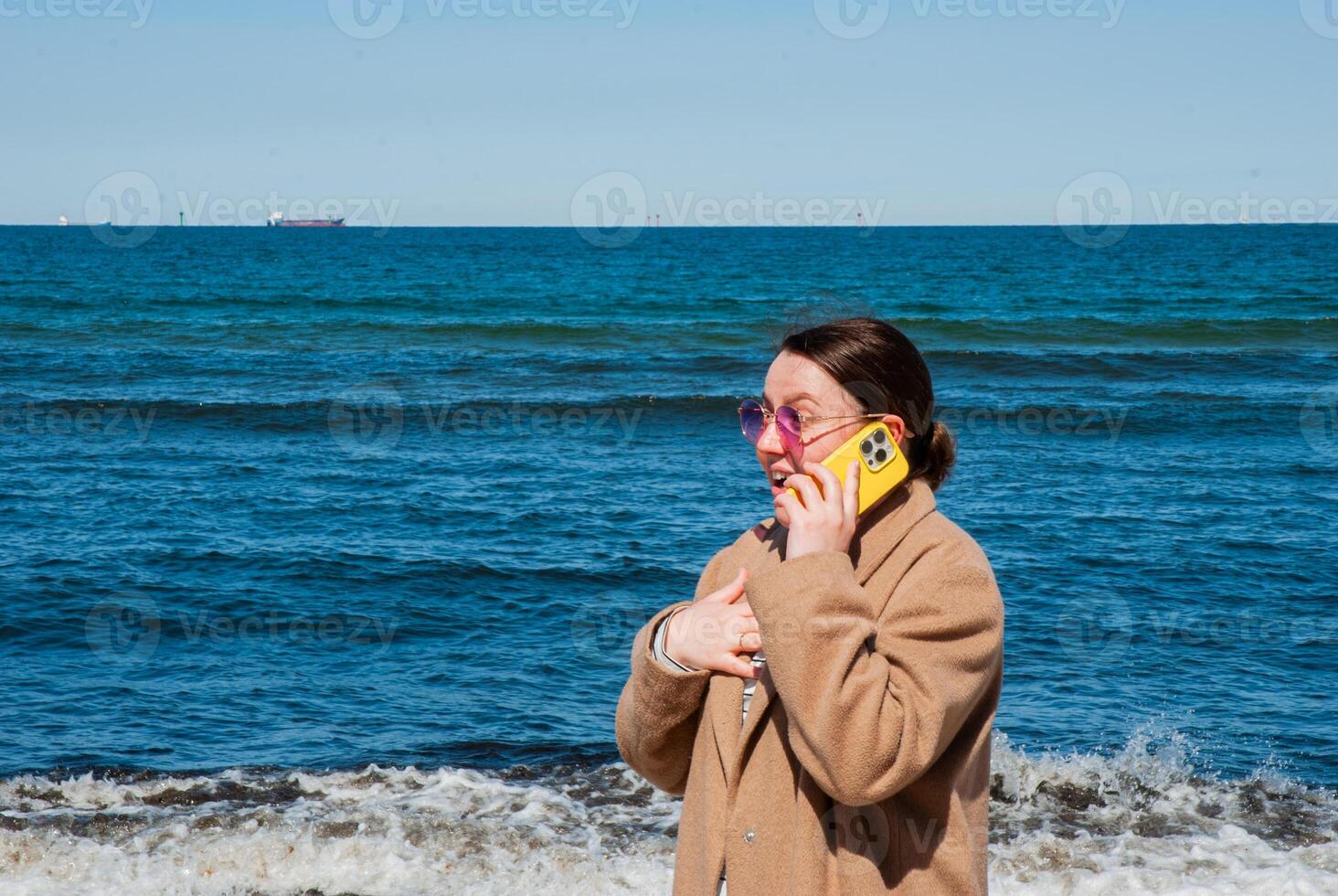 Young caucasian female at the beach wearing a brown coat, talking on a yellow phone with purple sunglasses, stands by the ocean on a clear day. Digital nomad lifestyle. Remote work photo