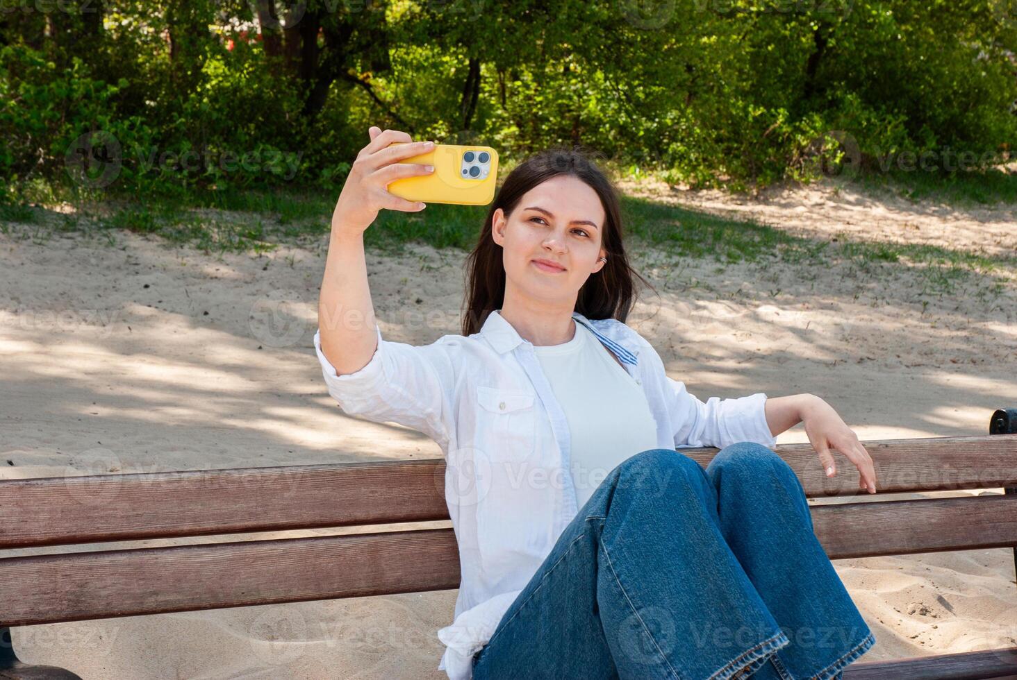Young caucasian woman taking a selfie with yellow smartphone on a sunny day at the beach, relaxing on wooden bench in casual outfit. Digital nomad lifestyle. Remote work photo