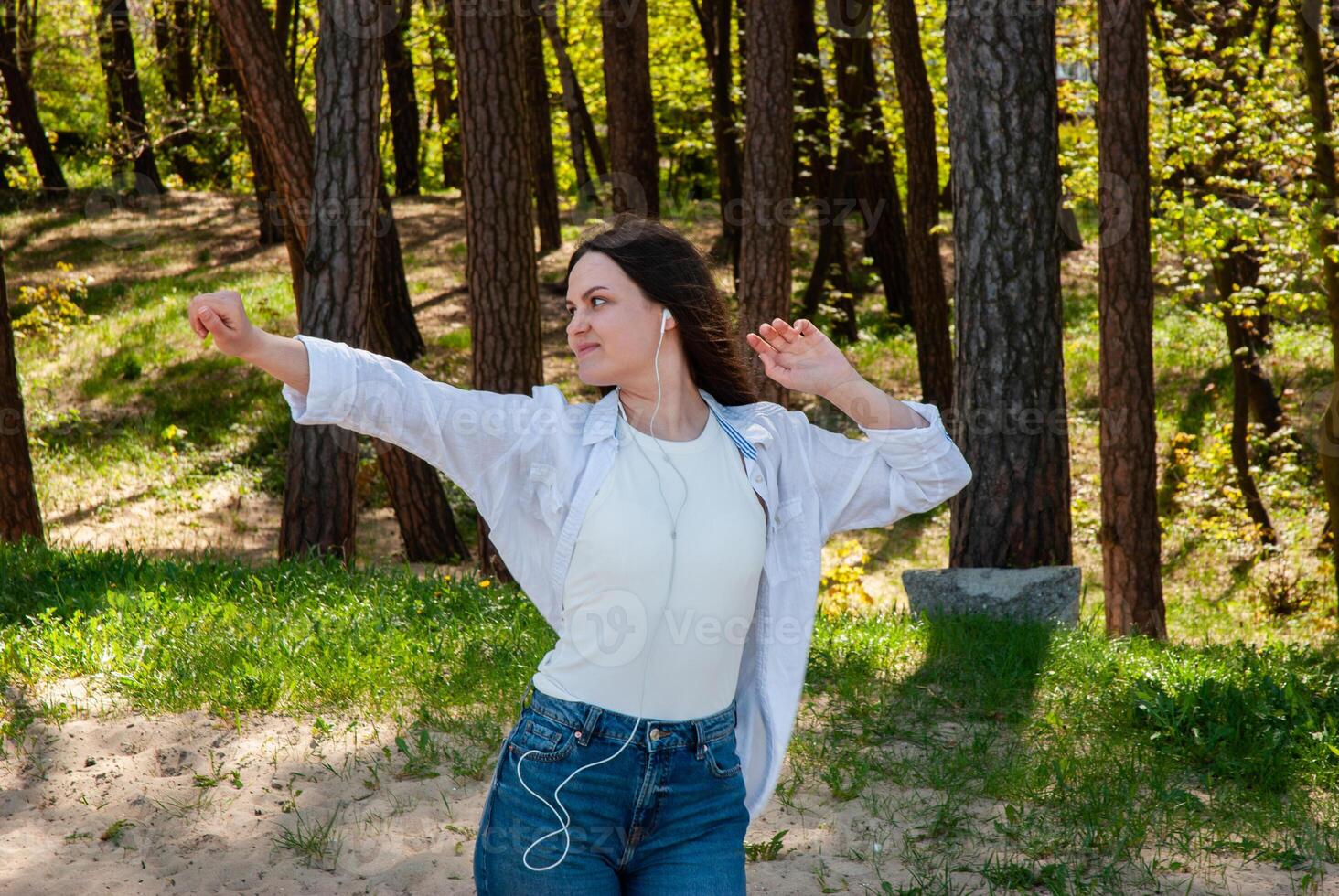 Young caucasian woman enjoying music while dancing in a sunlit forest, wearing casual clothing, expressing joy and freedom in nature. Digital nomad lifestyle. Remote work photo