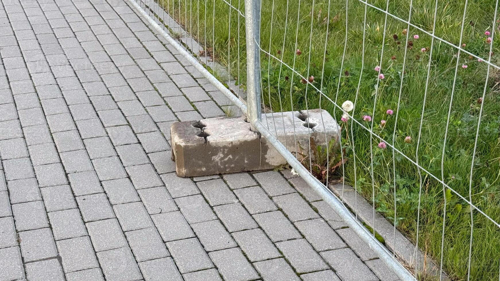 Sturdy grey concrete block secures metal fence on a varied texture pavement with green grass and pink flowers enhancing urban landscape photo