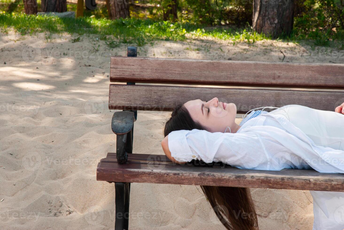 Young caucasian woman relaxing on wooden park bench at sandy beachside, enjoying peaceful summer afternoon under shade of trees. Digital nomad lifestyle. Remote work photo