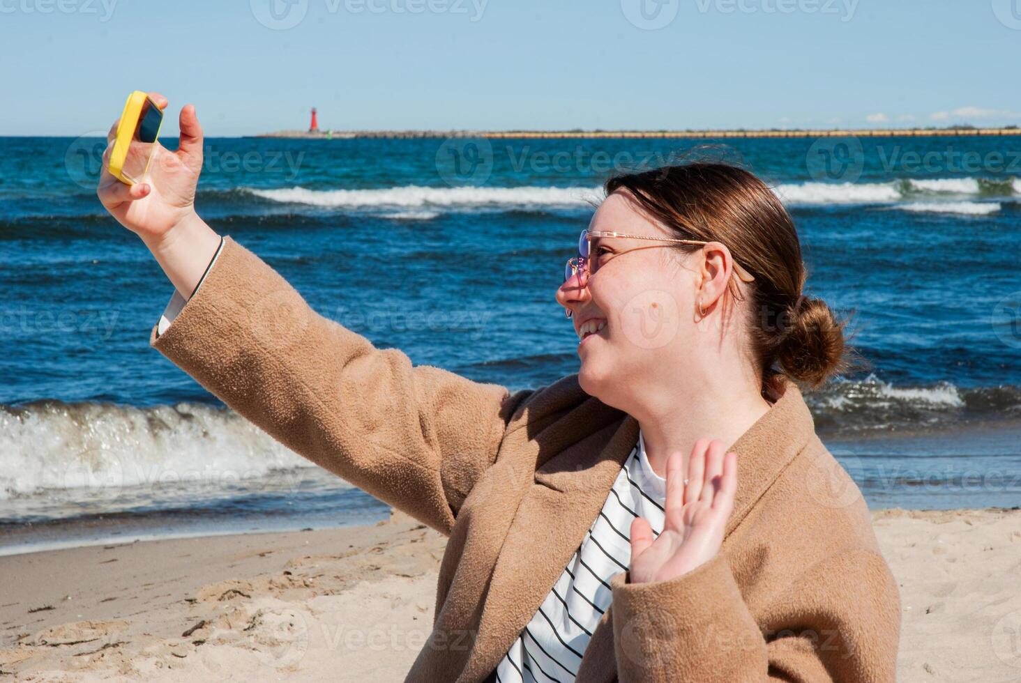 Caucasian young adult female capturing a selfie on a sunny beach day with waves and lighthouse in the background under clear blue sky. Digital nomad lifestyle. Remote work photo