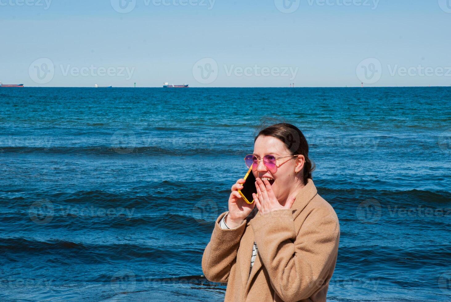 Young caucasian female adults joyfully talking on phone by ocean on a sunny day with clear blue skies and vibrant blue sea waves. Digital nomad lifestyle. Remote work photo
