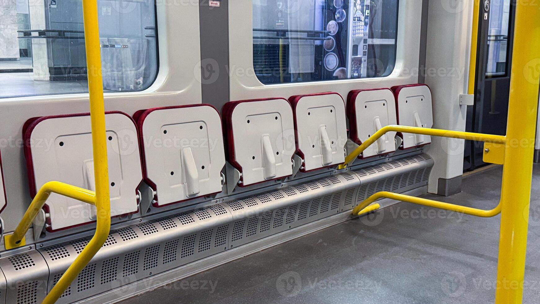 Modern urban subway interior featuring foldable red and white seats with bright yellow safety poles in a clean, spacious underground setting photo