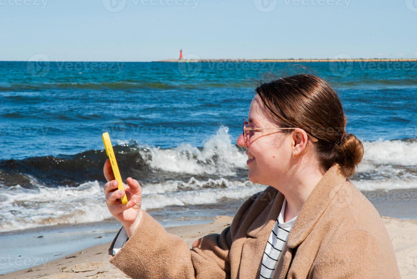 Young caucasian female enjoying a relaxing beach day, smiling as she takes a selfie with her phone against a backdrop of waves and a distant lighthouse. Digital nomad lifestyle. Remote work photo