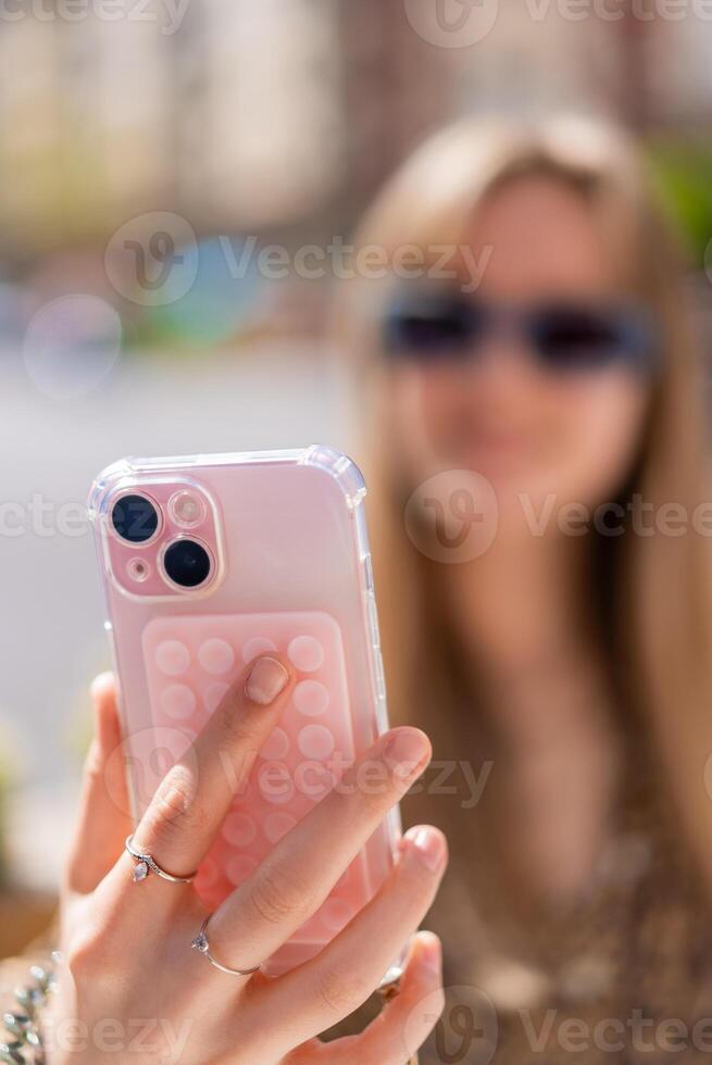 Close up of hands holding a smartphone with a pop it accessory and a transparent case, a young woman with sunglasses is blurred in the background, taking a selfie or recording a photo
