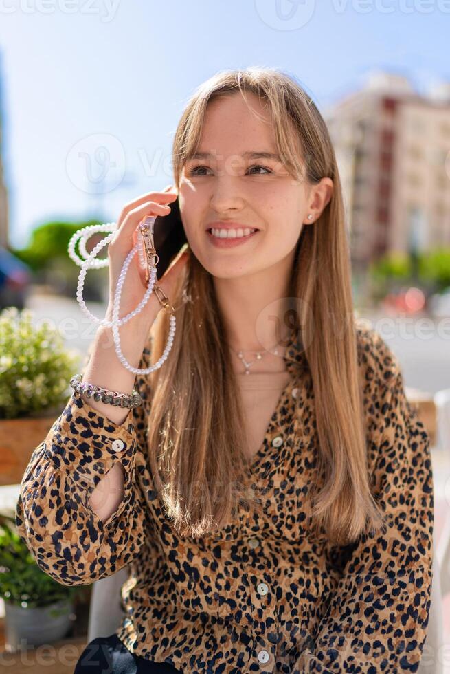 Young woman smiling and talking on a smartphone, showcasing a stylish beaded phone strap while enjoying the outdoors in a vibrant urban setting during a sunny day photo
