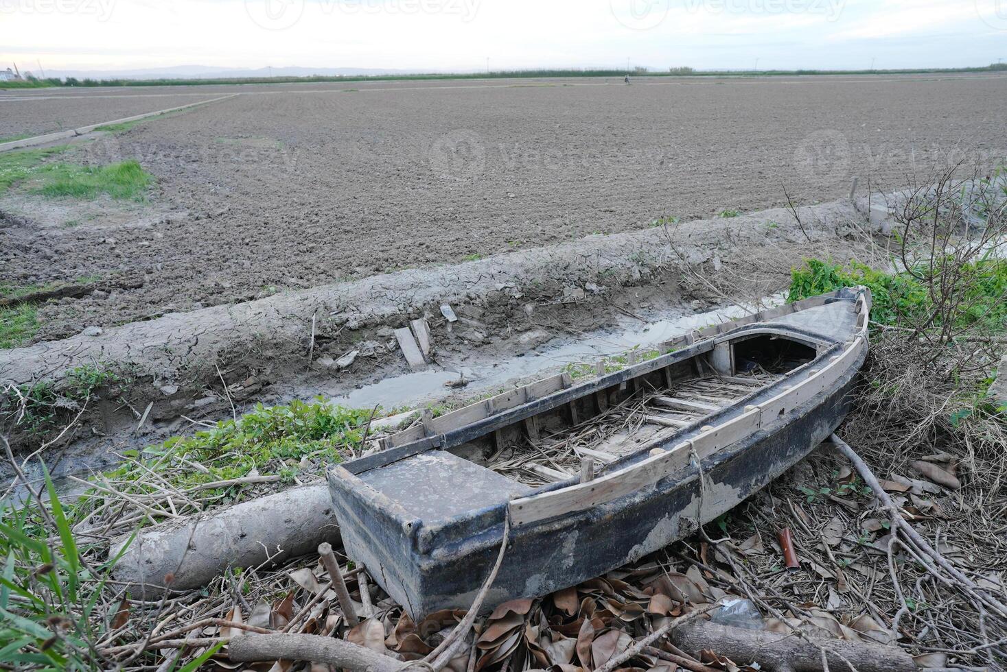 Abandoned old wooden boat lying in dry canal surrounded by grass mud and farmland under cloudy sky photo