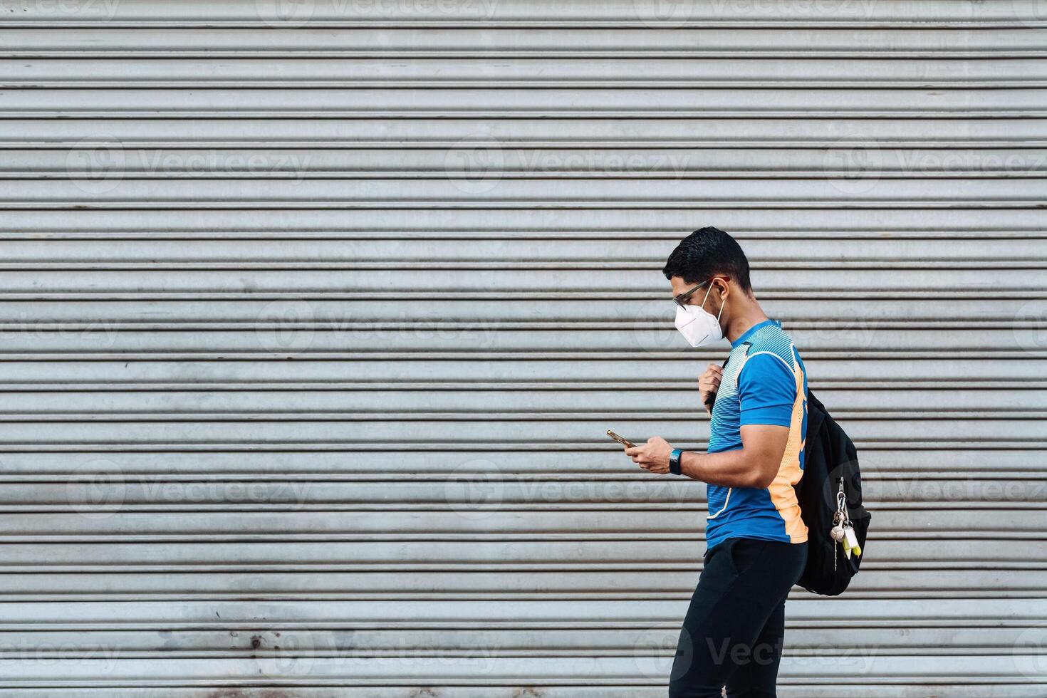 Hispanic male with a protective face mask and a backpack looking at his phone in front of a garage. photo