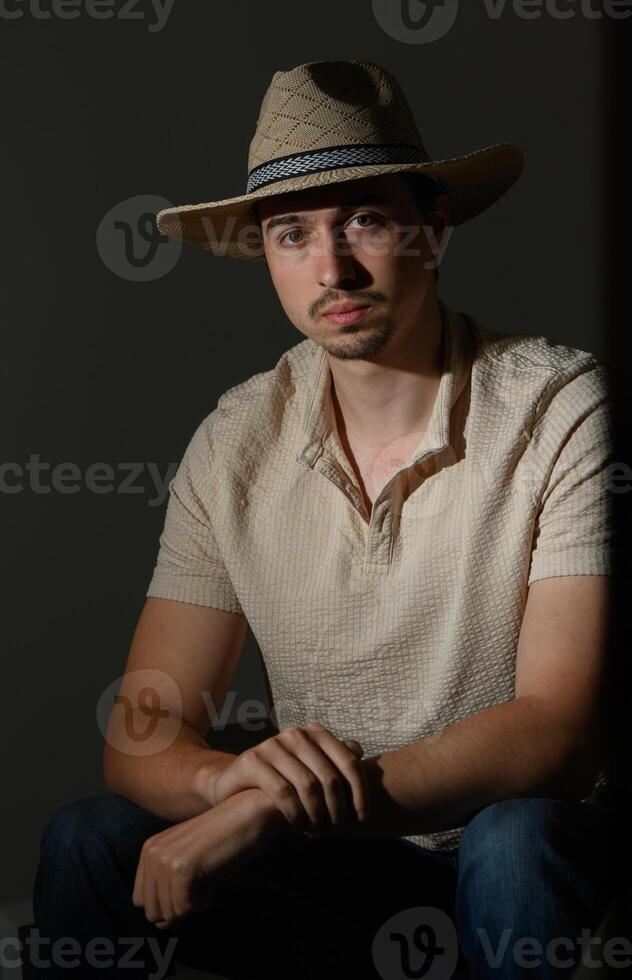 Portrait of a young man in a straw hat seated in dim lighting, reflecting thoughtful contemplation in a stylish setting photo