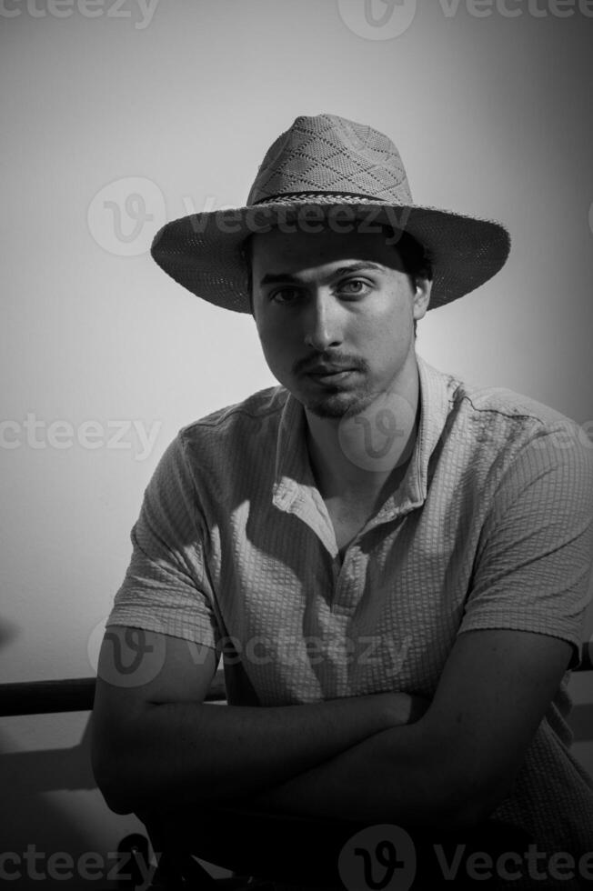 Man in a straw hat radiates calm confidence in monochrome setting while sitting in a casual pose photo