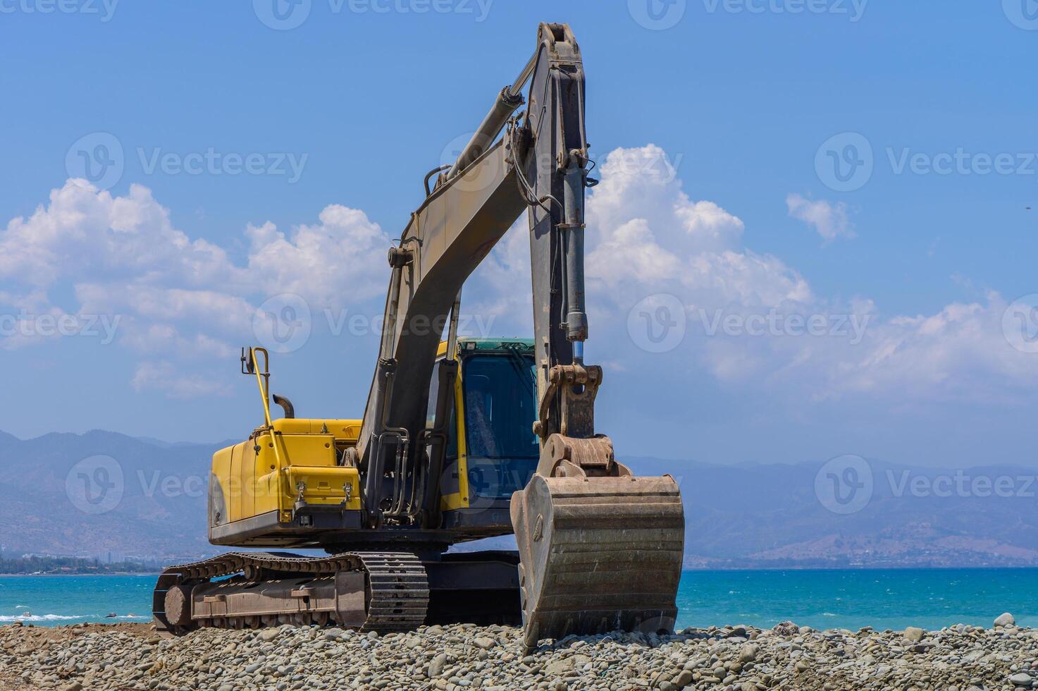Yellow excavator operating on a Mediterranean beach in Cyprus, leveling sand during coastal maintenance. Clear sky, sea in background. photo