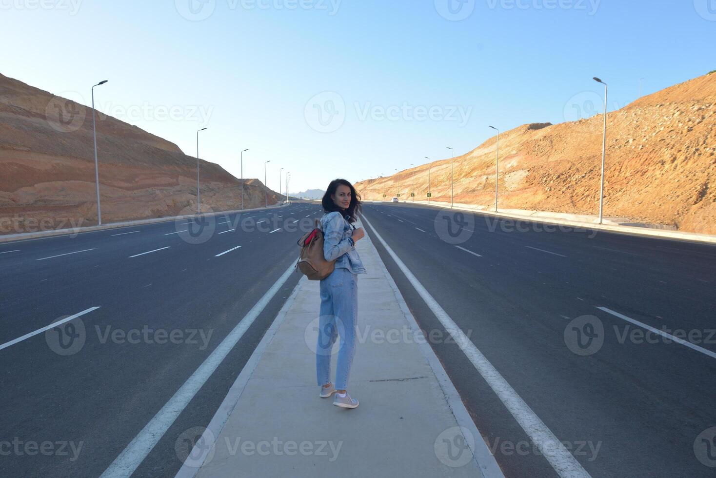 A girl in a denim suit with a backpack walks in the center of a quiet road among the mountains at sunset. photo