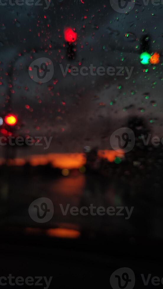 Blurred view through rain-soaked windshield capturing traffic lights glowing against dark stormy sky with hints of sunset creating dramatic urban atmosphere photo