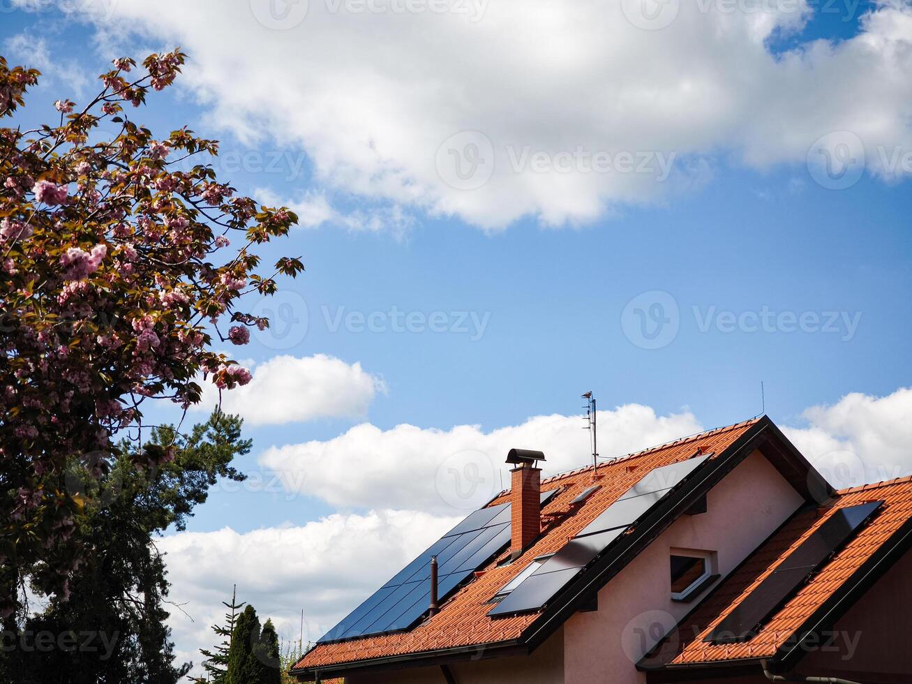 Solar panels installed on a modern house roof surrounded by blooming trees under a bright blue sky with fluffy clouds showcasing renewable energy concept photo