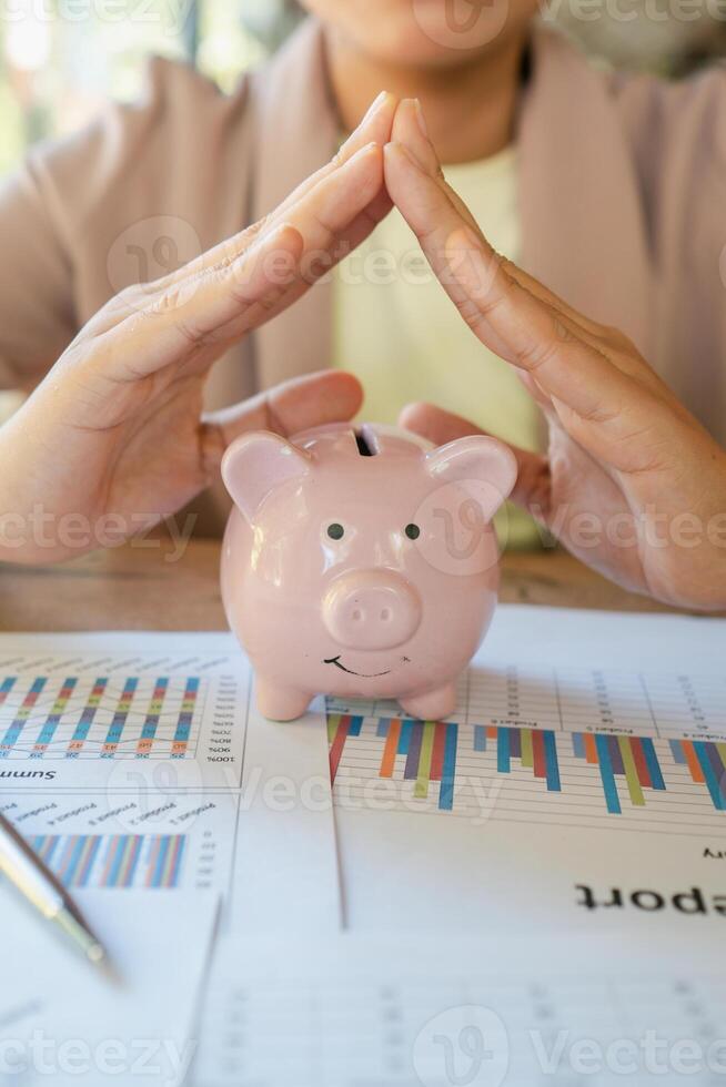 Close-up of hands forming a roof over a pink piggy bank on financial documents with graphs, symbolizing savings and financial security. photo