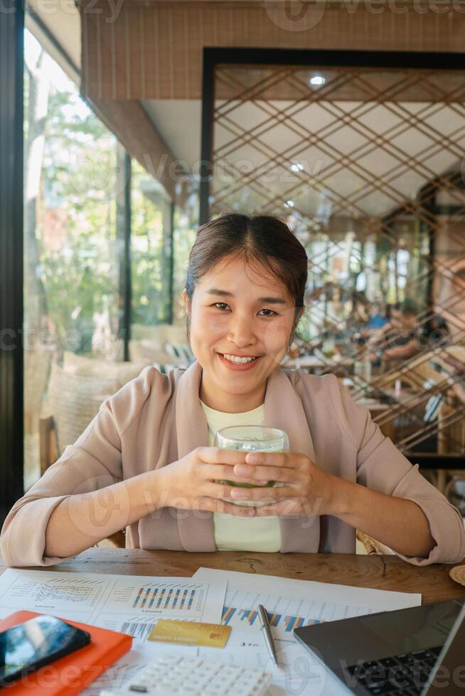 Smiling woman holding coffee cup in a modern cafe setting, surrounded by documents and a laptop, showcasing a work-life balance. photo
