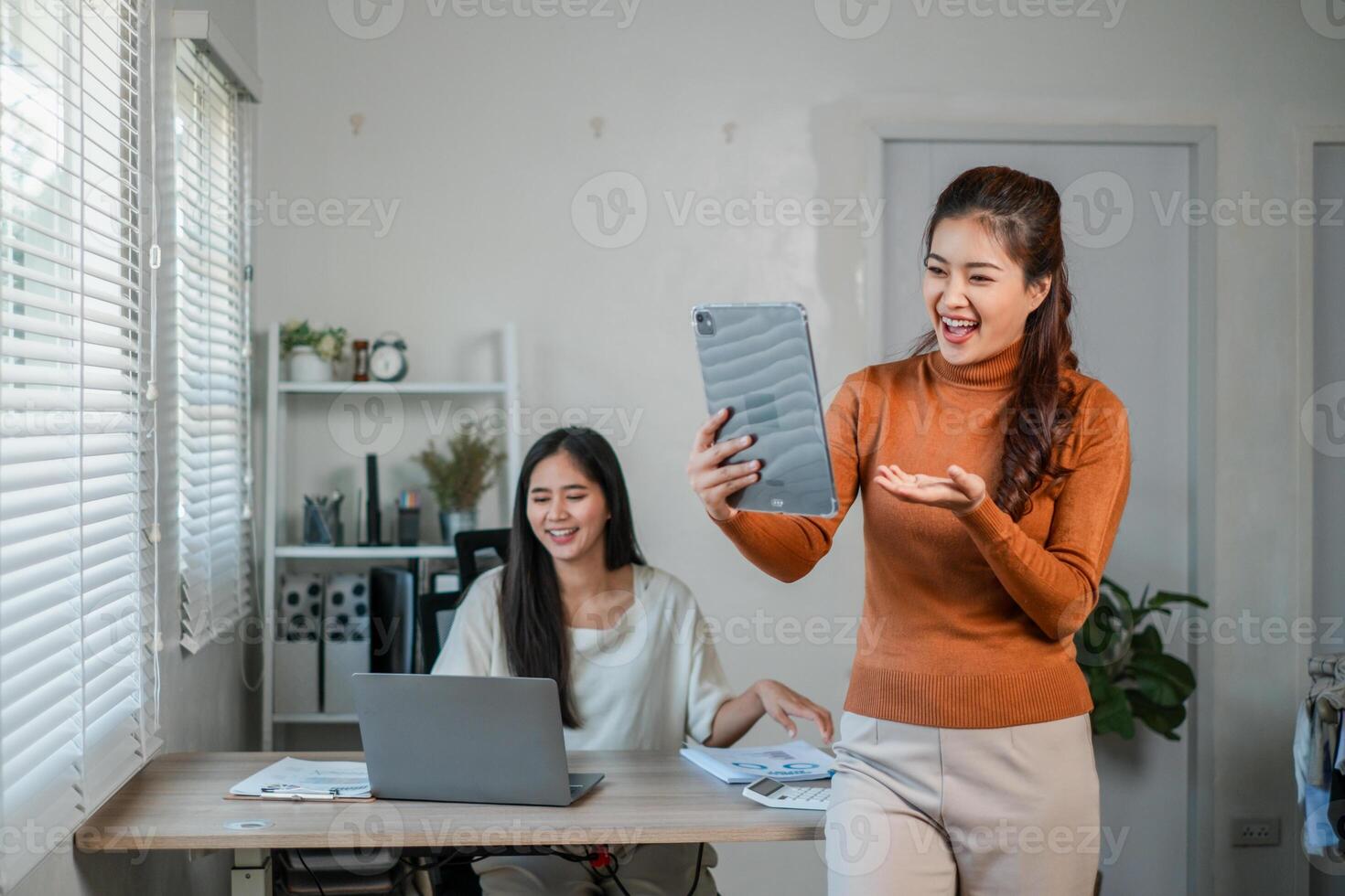 Two women in a bright office, using laptops and a tablet, engaged in a call and collaborative work. photo