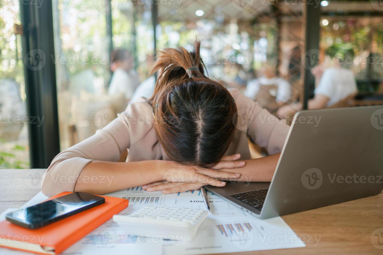 Tired woman resting her head on a desk surrounded by a laptop, documents, and a smartphone in a sunlit office. photo