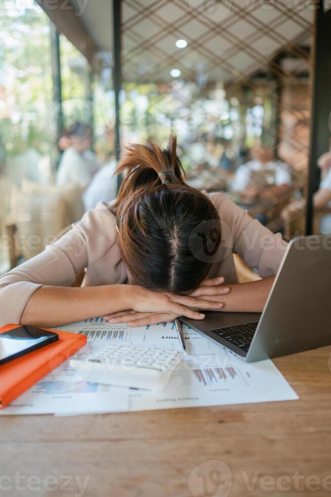 Tired businesswoman with head on desk surrounded by documents, laptop, and smartphone in a busy office environment. photo