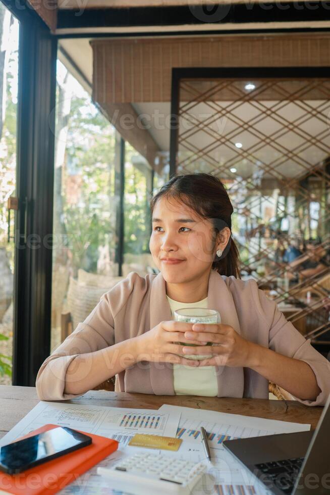 Smiling businesswoman holding coffee cup, sitting at desk with documents and laptop, in a bright, modern office space. photo