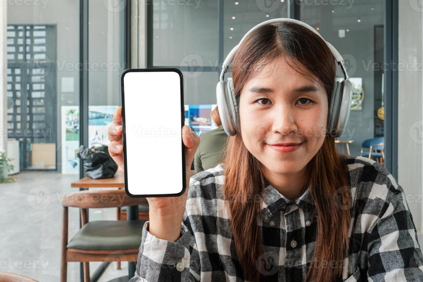 Smiling woman wearing headphones displays a smartphone with a blank screen in a contemporary cafe environment. photo