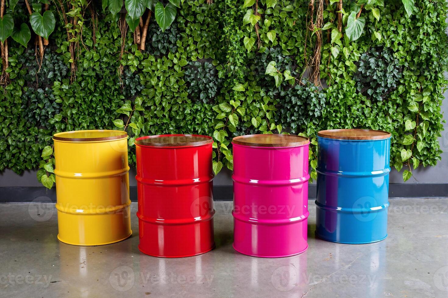 three brightly colored metal oil drums in yellow, pink, and blue, standing in a row against a lush green vertical garden wall. Striking contrast between industrial objects and vibrant nature design photo