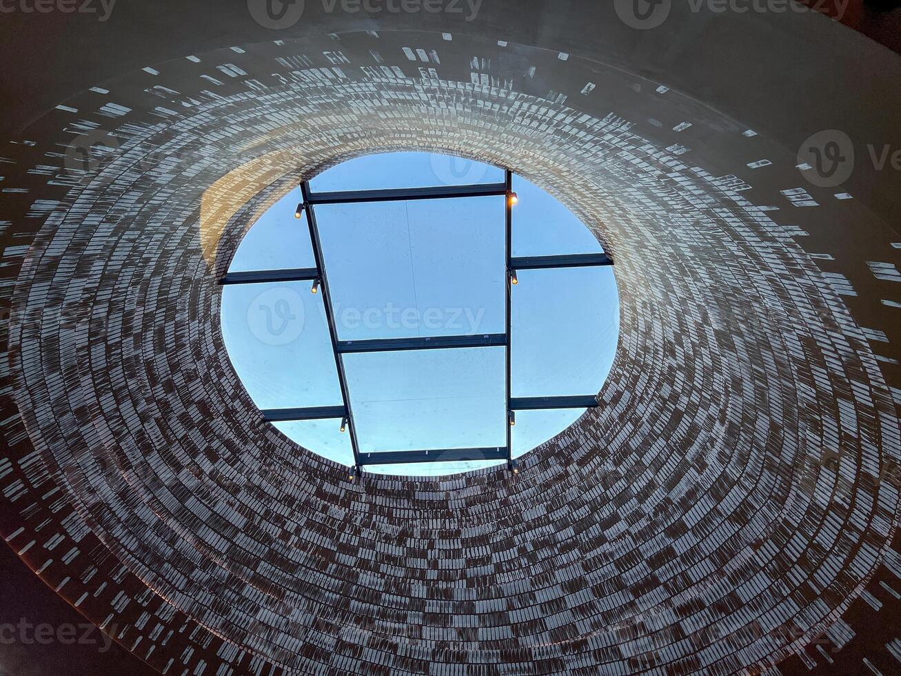Gazing up at the expansive sky through a beautifully designed round window with textured walls, a circular opening reveals a bright blue sky crossed by dark beams photo