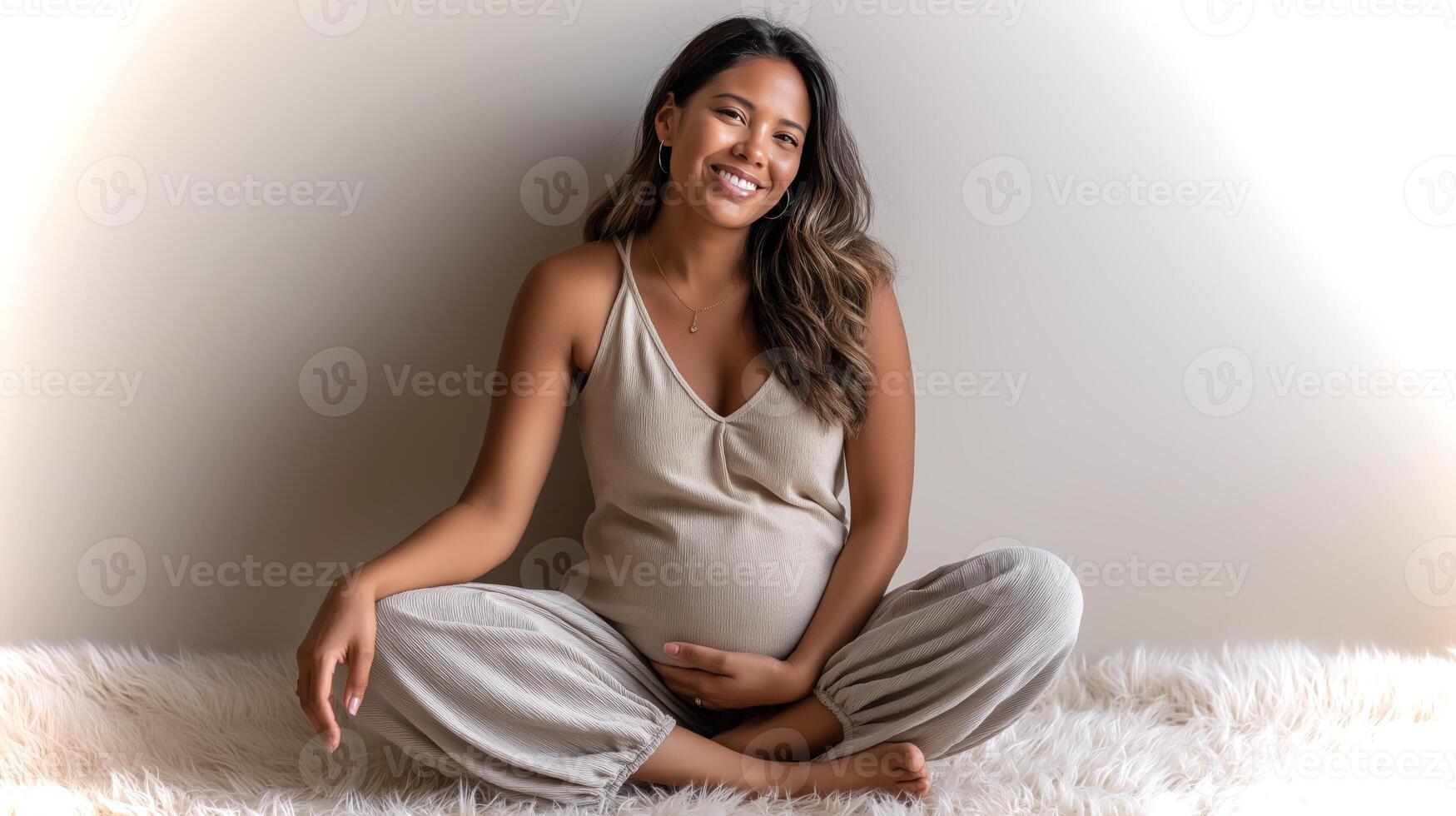 A latina woman with a glowing smile sits comfortably on a soft rug, showcasing her pregnancy while wearing a beige tank top and loose flowy pants. photo