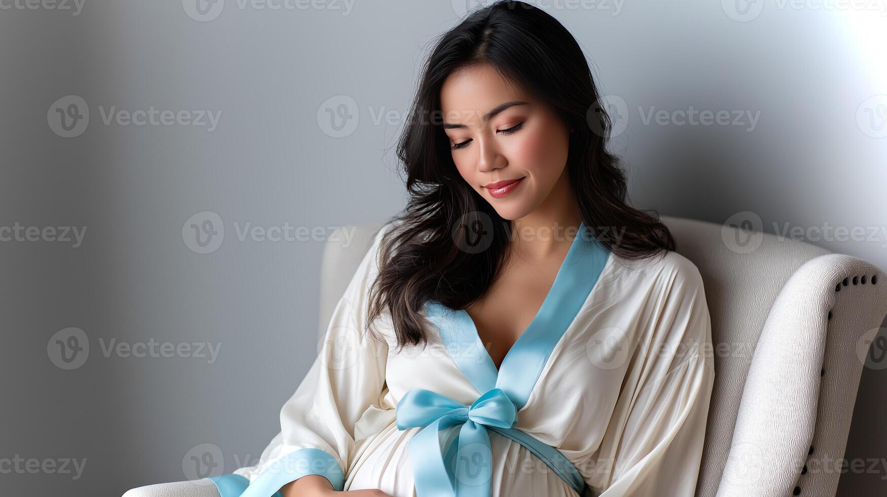 A serene moment captures a pregnant woman in a cream robe with a light blue sash, seated comfortably and embracing her journey into motherhood. photo