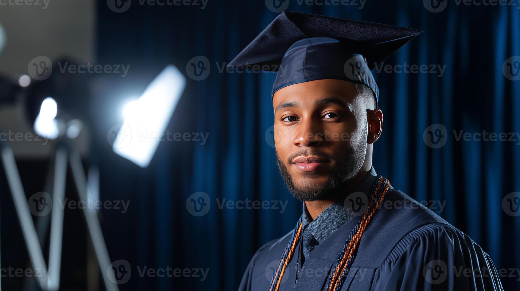 A graduate stands proudly holding his diploma, wearing a cap and gown, as he poses for an official photo in a formal setting.