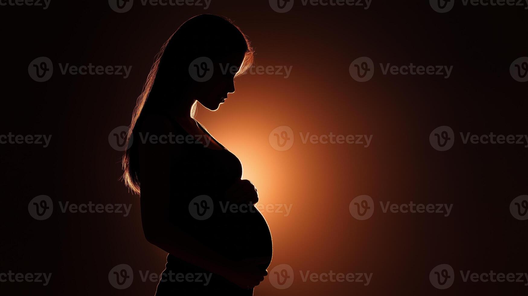A pregnant woman stands in silhouette, supporting her belly gently with her hands while illuminated by warm, soft light in the background. photo