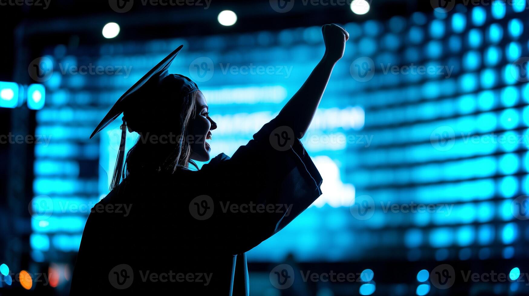 A proud salutatorian stands with a raised fist, celebrating a significant achievement at a graduation ceremony, illuminated by a vibrant blue backdrop. photo