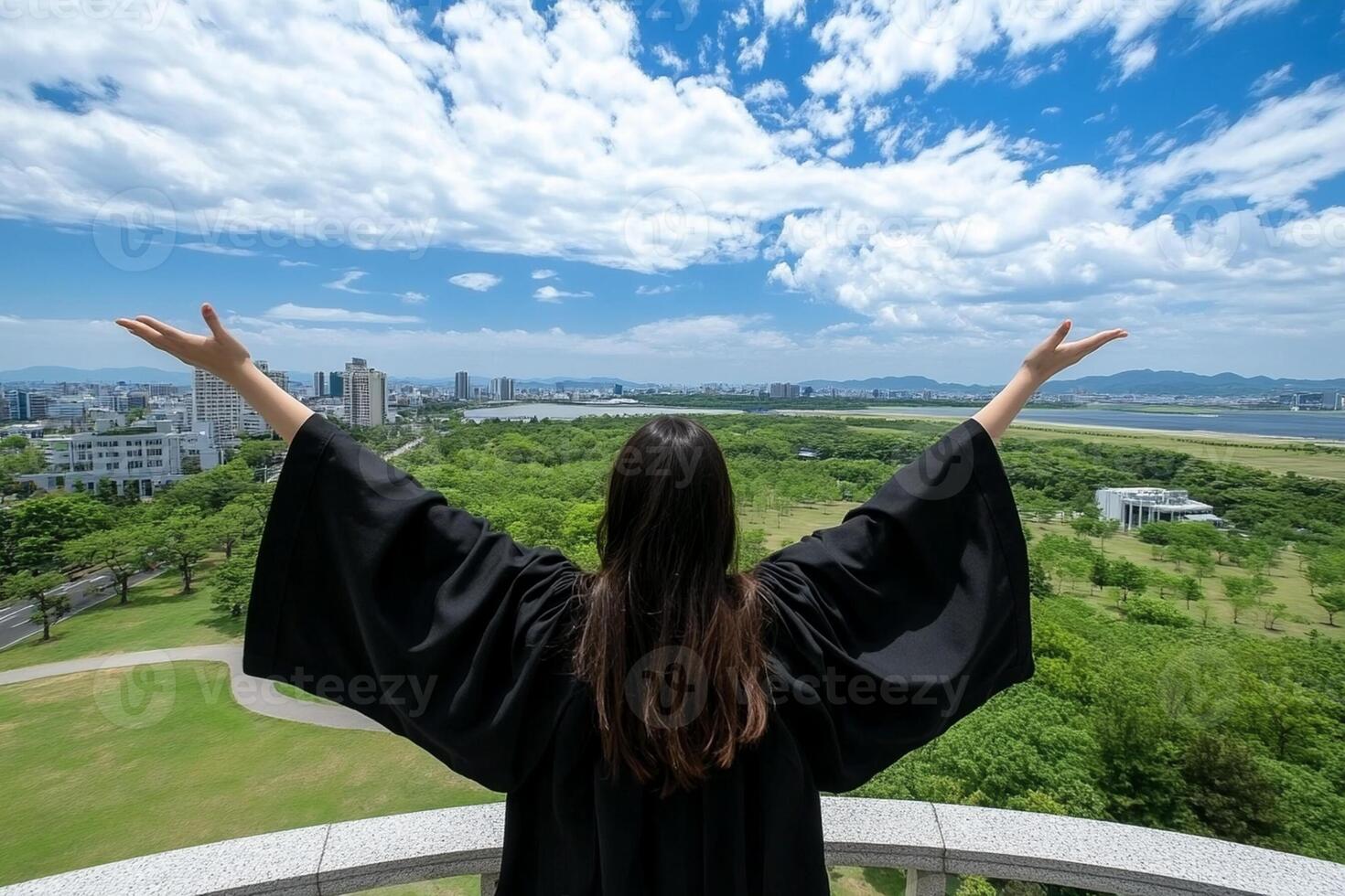 A woman in a graduation gown stands on a balcony overlooking a city photo