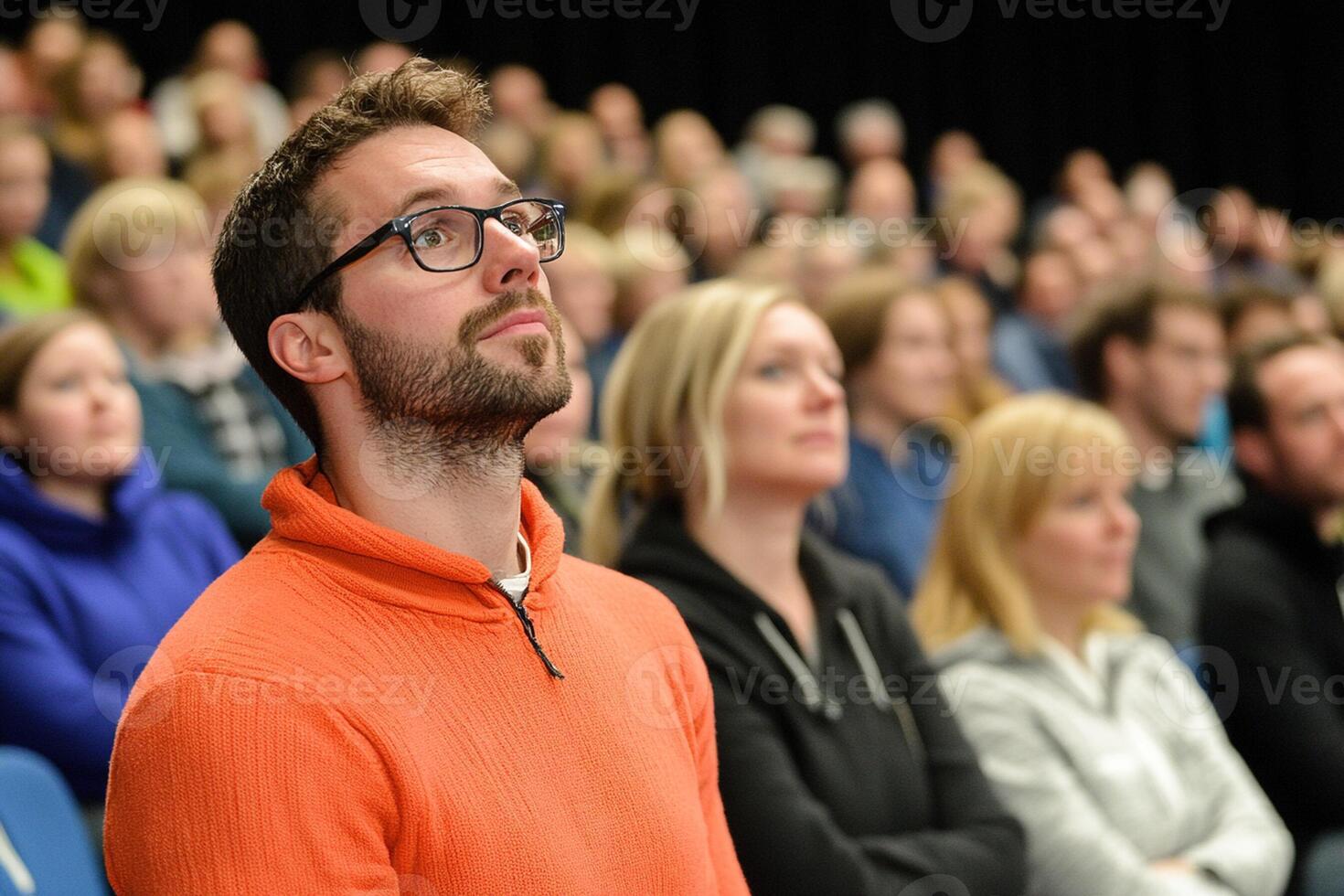 A man with glasses and an orange sweater is sitting in front of a crowd photo
