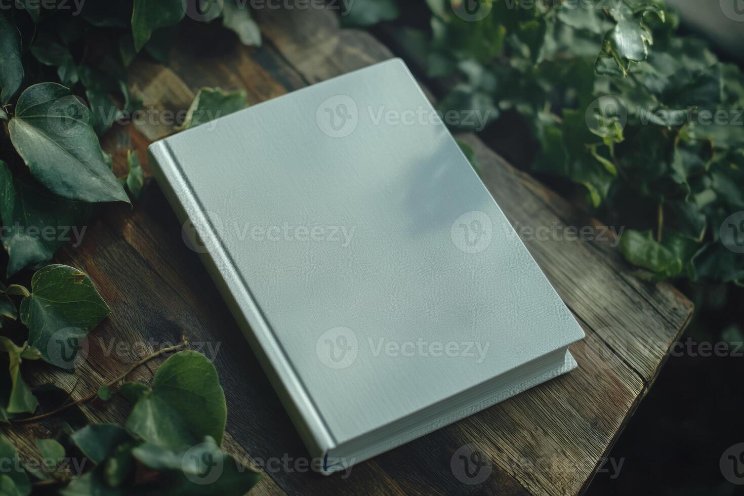 Minimalist mockup of a blank book placed on a rustic wooden table surrounded by lush green leaves in soft, diffused light photo
