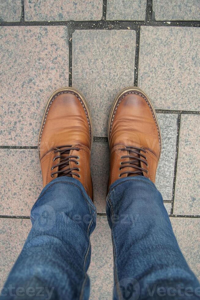 A casual male outfit featuring denim jeans and brown leather shoes while standing on a textured pavement during the day photo
